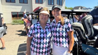 Two golf tournament participants in matching argyle shirts pose beside golf carts outside the clubhouse at the Community Health Foundation of Wright County Golf Tournament.