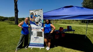 Two Home Energy Heating & Cooling representatives stand beside a sponsor tent and company banner at the Community Health Foundation of Wright County Golf Tournament.