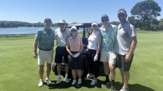 Six golf tournament participants pose beside a golf cart on the course with a lake in the background at the Community Health Foundation of Wright County Golf Tournament.